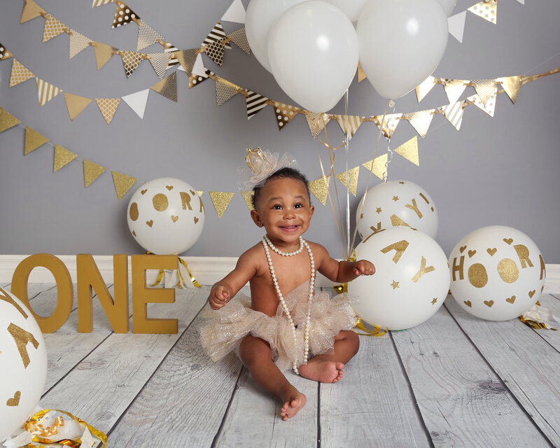 A smiling baby girl in a gold tutu with pearl accessories, sitting with gold balloons and bunting in a glamorous cake smash setup.