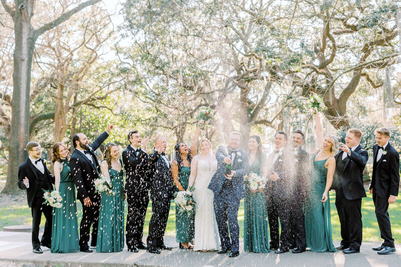 Bridal party standing outside with groom opening bottle of champagne