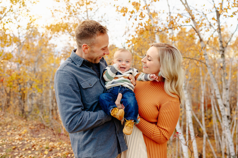 A smiling couple stands in an autumn forest, holding a cheerful baby dressed in a striped sweater and jeans. The golden leaves create a warm and cozy atmosphere, beautifully captured by a talented Minnesota photographer specializing in Brainerd Lakes photography.