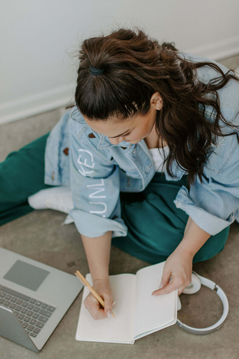 woman with dark hair sitting on floor in jean jacket teal pants journaling with her laptop and notebook
