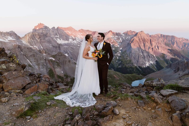 Bride and groom standing on rocky summit of Mount Sneffels at sunrise with dramatic alpine peaks and a glacial lake in the background during their July elopement in Telluride, Colorado