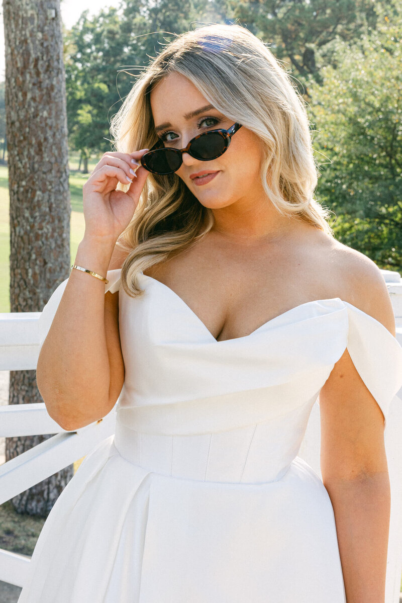 a wedding photo of a bride in a white dress holding a pair of sunglasses in her hand as she gently looks at the camera