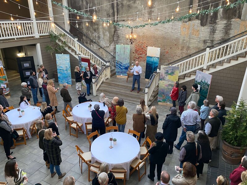 A man stands on stairs speaking to a gathered crowd at an art exhibit on the stairs in an open couryard. Large paintings are displayed around him, and people listen and observe from below.