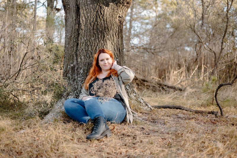 Photo of photographer Bethany Simms sitting agains a tree wearing a green shirt and jeans with black books