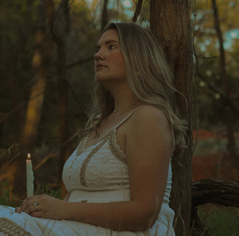 A woman is sitting and leaning against a tree as she is sitting down. She is looking away from the camera and is holding a candle stick.
