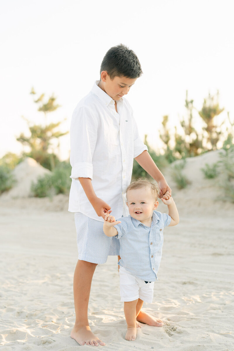 older brother in white shirt and seasucker shorts holds baby brother's hands at the beach in Delaware