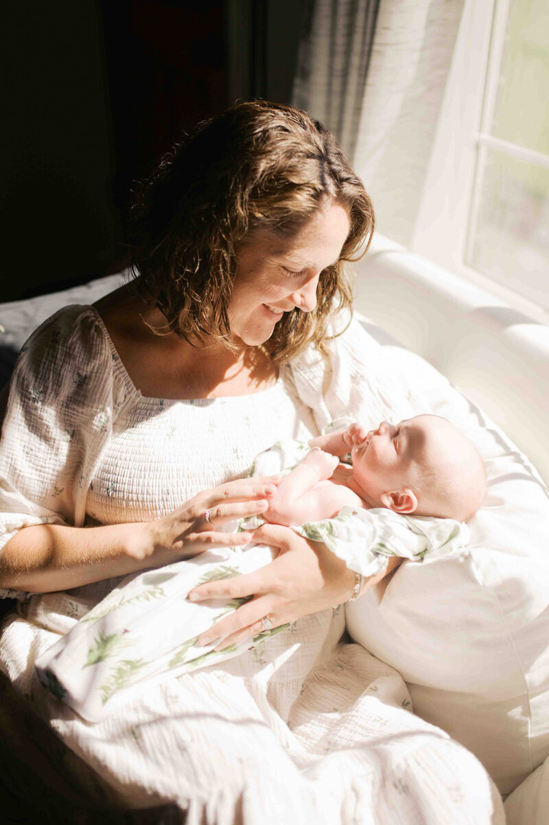 Mom sits on white sofa with sunshine shining on her face as she smiles down at newborn loosely wrapped in swaddle by Portland Family Photographer Emilie Phillipson Photography. 