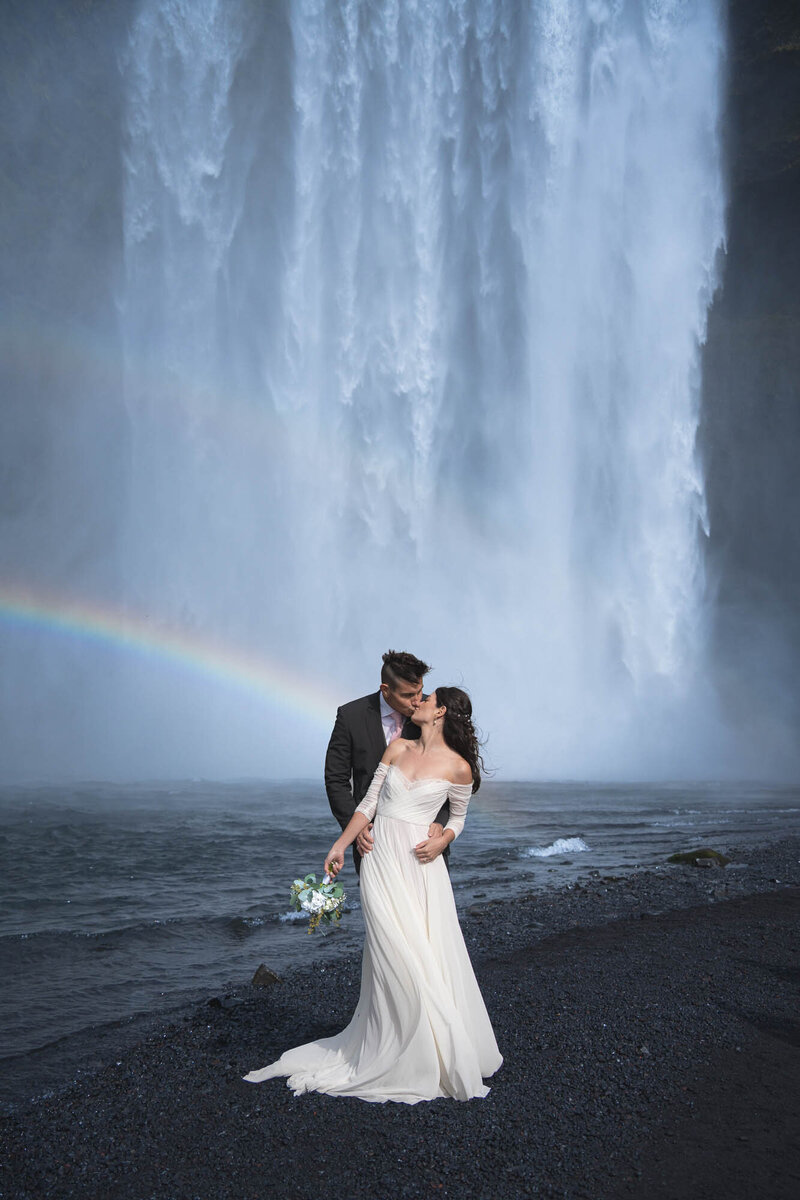 Couple in their wedding attire kissing front of Skogafoss in iceland with a rainbow behind them.