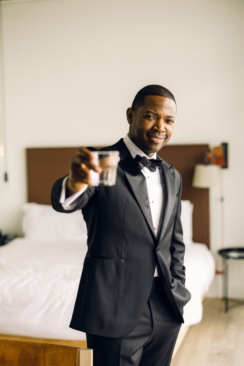 A groom, dressed in a black tux and bow tie, looking at the camera to "cheers" with a glass of whiskey.