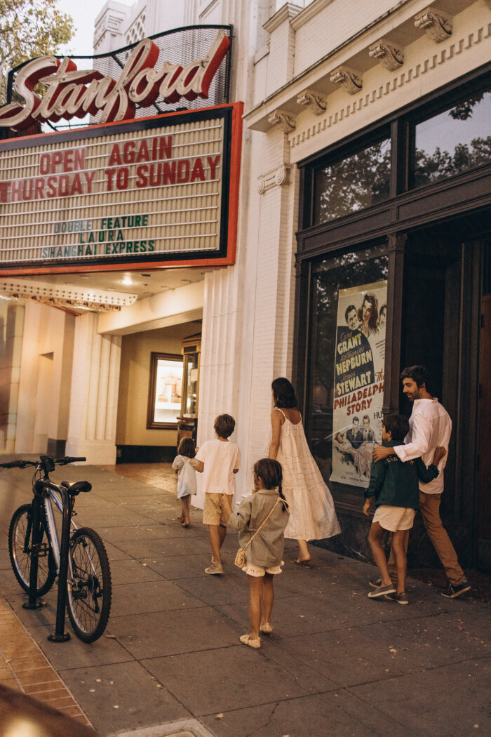 A family of six, dressed semi-formally, walks towards the entrance of the stanford theater with movie posters displayed, under a lit marquee announcing a re-opening.