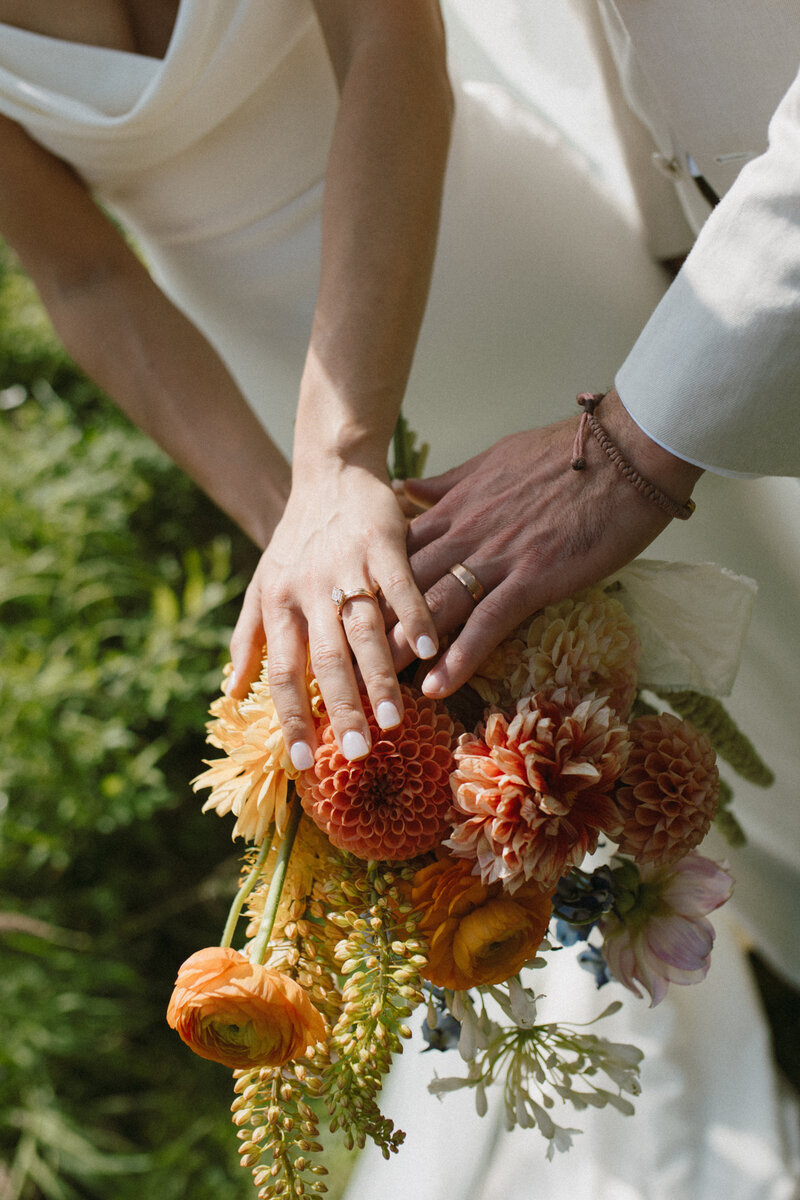bride and groom place hands with wedding rings over wedding bouquet at alberta wedding