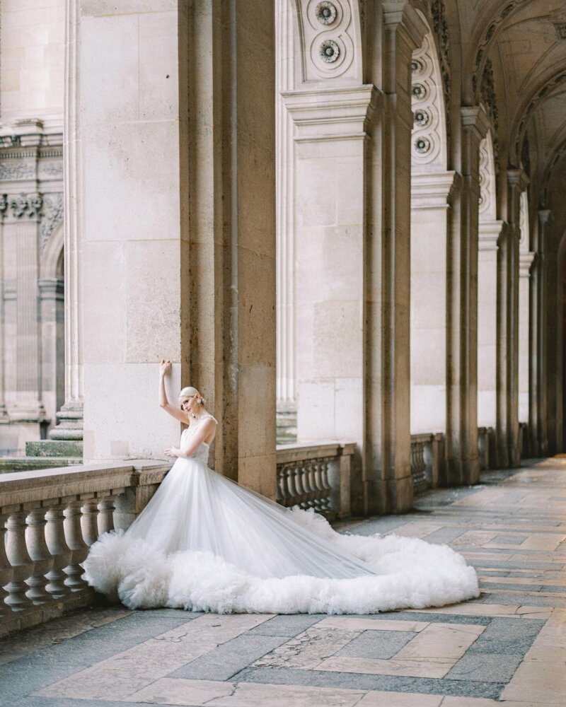 A bride in a voluminous white wedding dress poses gracefully against a stone railing in an elegant, arch-filled corridor in the Louvre, Paris. The dramatic gown trails behind her on the checkered floor, its beauty perfectly captured by luxury international wedding photographer Andreas K. Georgiou.