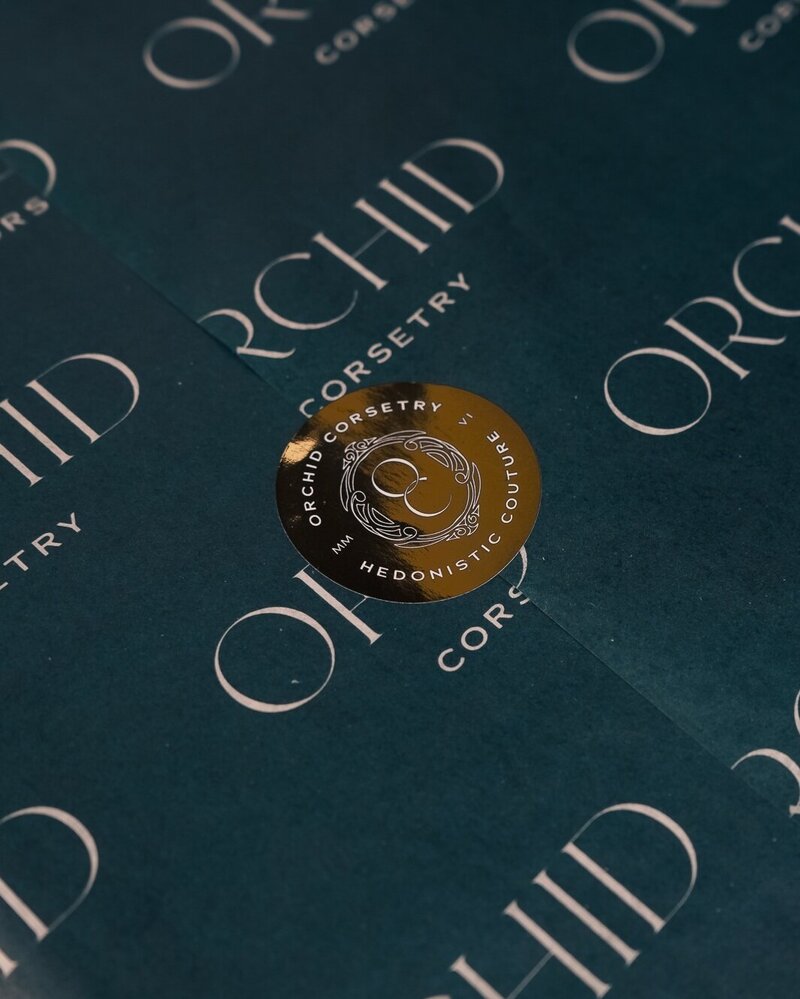 Branded gold sticker featuring Orchid Corsetry’s emblem on teal tissue wrap