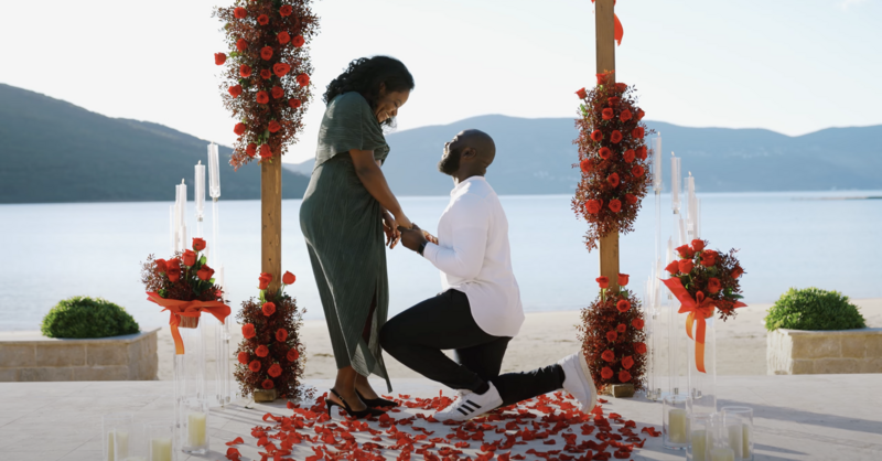 Man on one knee proposing to his partner under a red floral arch by the water, during a heartfelt and cinematic surprise proposal video in Montenegro