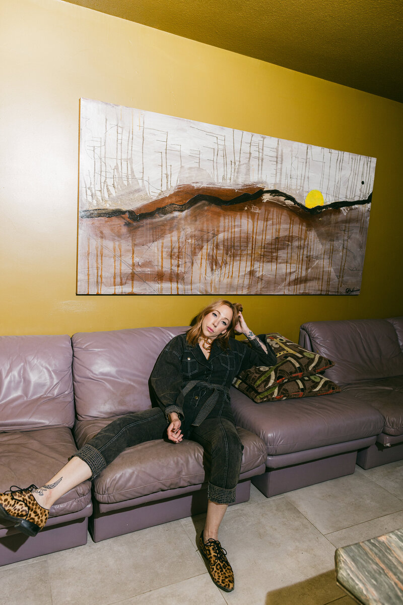 Bold branding image of a woman lounging on a lavender couch beneath abstract wall art, styled in black denim, photographed in Greenville SC by Dani Lacey Photographs.