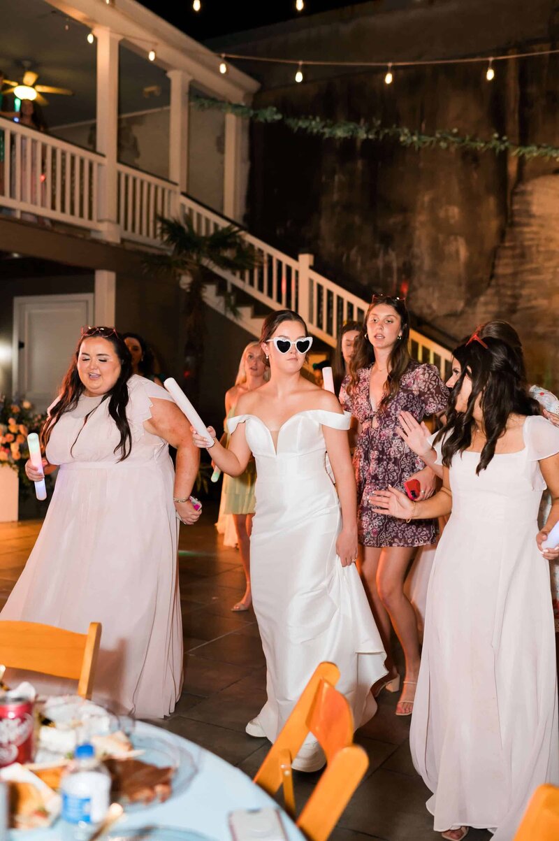 A bride in a white gown and heart-shaped sunglasses walks with bridesmaids in light pink dresses, holding glow sticks, at an outdoor wedding reception with string lights and a staircase in the background.