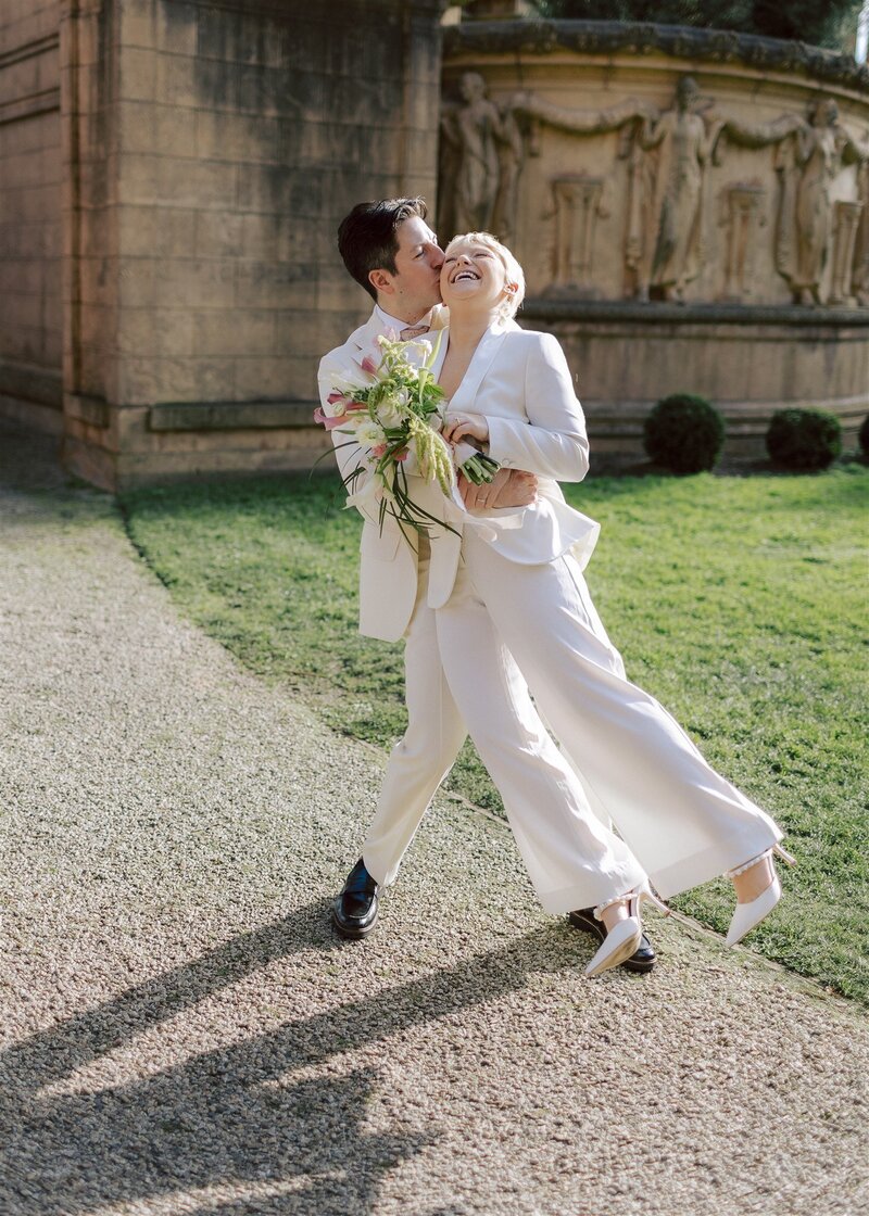 Non-binary couple in white suits holding bouquets, one lifts the other up playfully, lgbtq