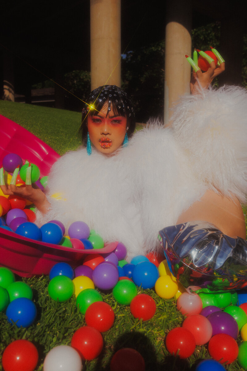 An asian woman wearing bright red sunglasses and a white top laying on a hammock filled with bright colored balls