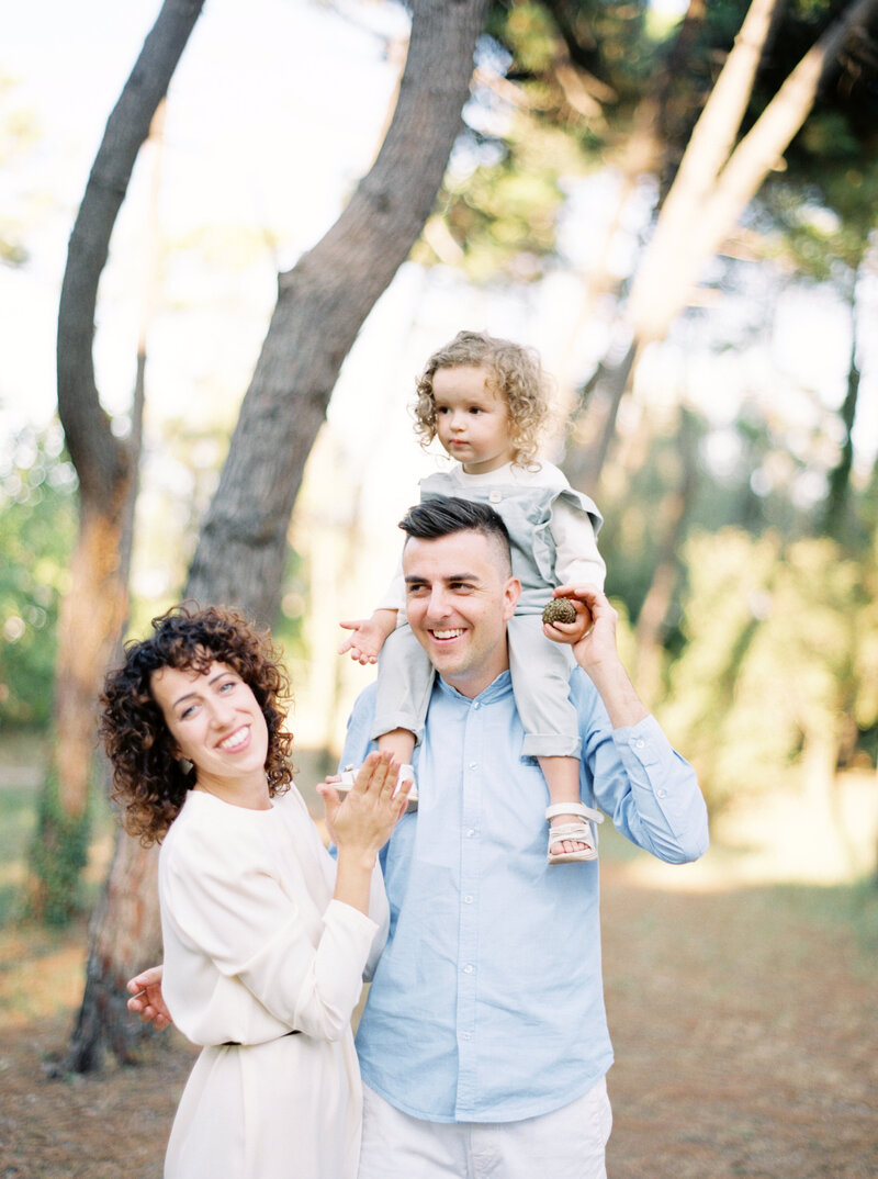 Little girl sits on her dad's shoulders with mom smiling standing with them