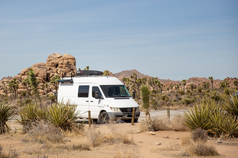 White Sprinter van parked among Joshua trees and desert rock formations in Joshua Tree National Park.