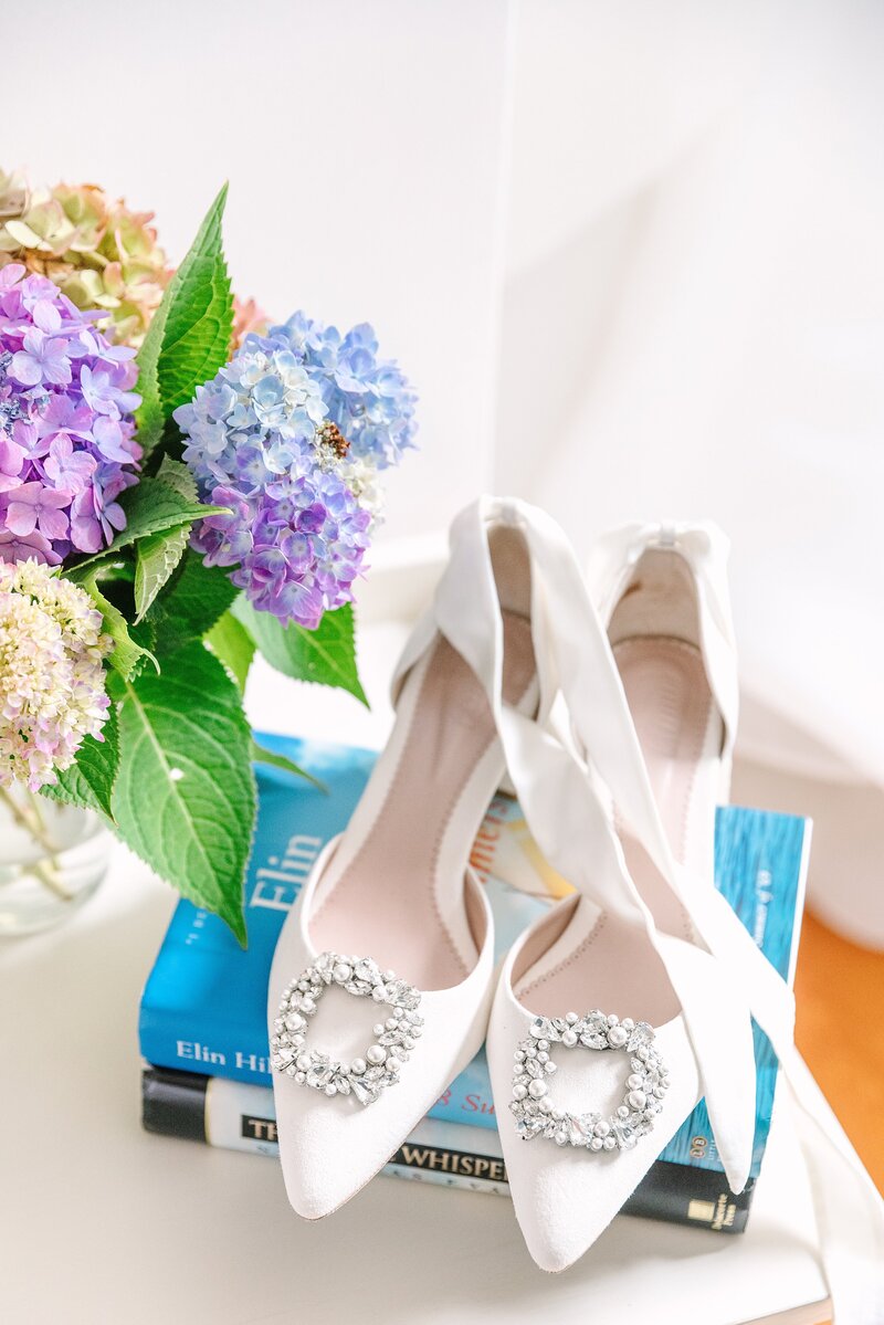 white shoes with ribbon tie ankle straps sitting on nantucket books next to blue and purple hydrangea