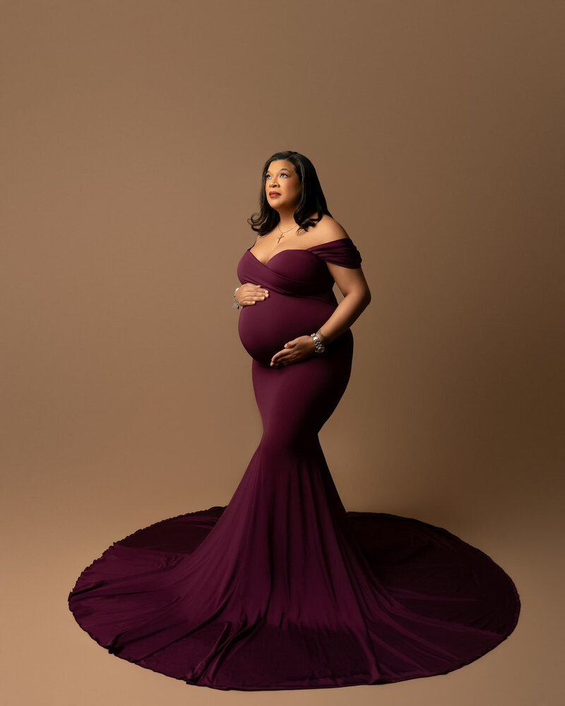 Studio maternity photo featuring deep plum fitted dress with off-the-shoulder neckline and dramatic silhouette