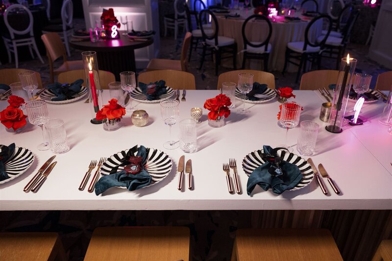Aerial view of a modern gothic tablescape in the ballroom of the Line Hotel DC of a white table with black and white striped dinner plates with a teal velvet napkin on top with black and hot pink alternating heart shaped placecards, white and black striped handle flatware, and ribbed modern glassware with hot pink floral with white bud vases and hot pink taper candles