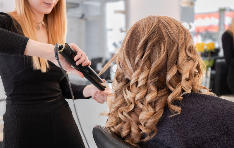 Frau mit Balayage sitzt auf dem Stuhl, Friseur lockt ihr Haar