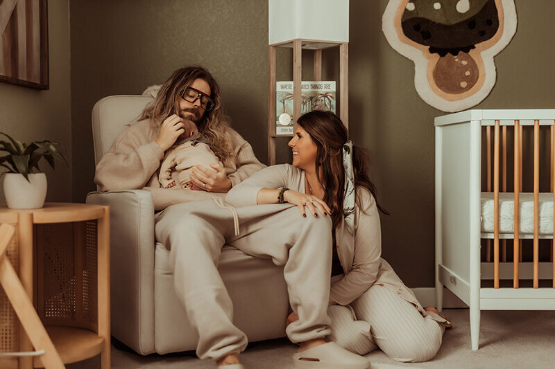 Dad sitting in a rocking chair holding a newborn baby boy while mom sits on the ground resting on dads knee