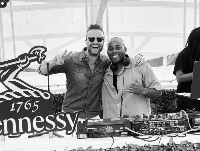 Black-and-white photo of two smiling DJs posing behind their decks at a Hennessy-branded outdoor event.