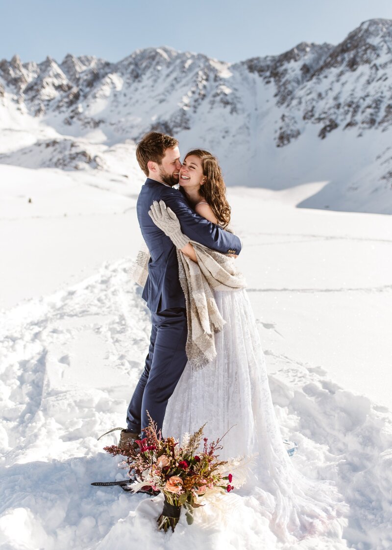 Colorado snowshoeing elopement photographer captures a couple in wedding attire embracing at Mayflower Gulch with snow-covered peaks in the background.