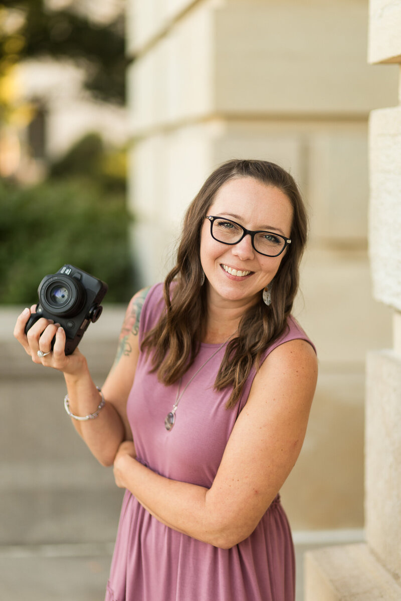 Brunette female in a purple dress wearing black glasses holding a black camera and smiling