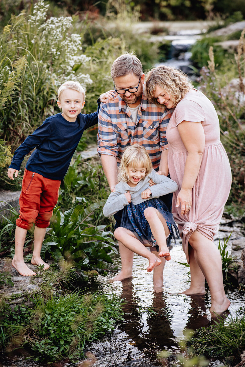 A family of four gathers at a small creek, with the father gently lifting a young girl into the water while the mother and older brother smile and watch.