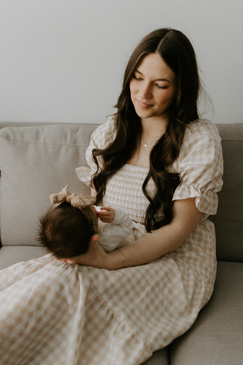 A mom sits on a sofa while holding and looking at her newborn baby.