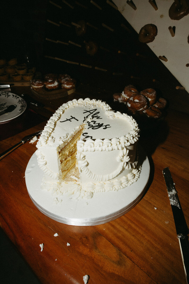 A white-frosted cake adorned with floral decorations sits on a wooden table, capturing the essence of romance. With a slice cut out, the top delightfully reads "The Firsts." Plates and pastries in the background complete this scene from a wedding at The Farmhill in Peterborough captured by Toronto wedding photographer Jennifer van Son.