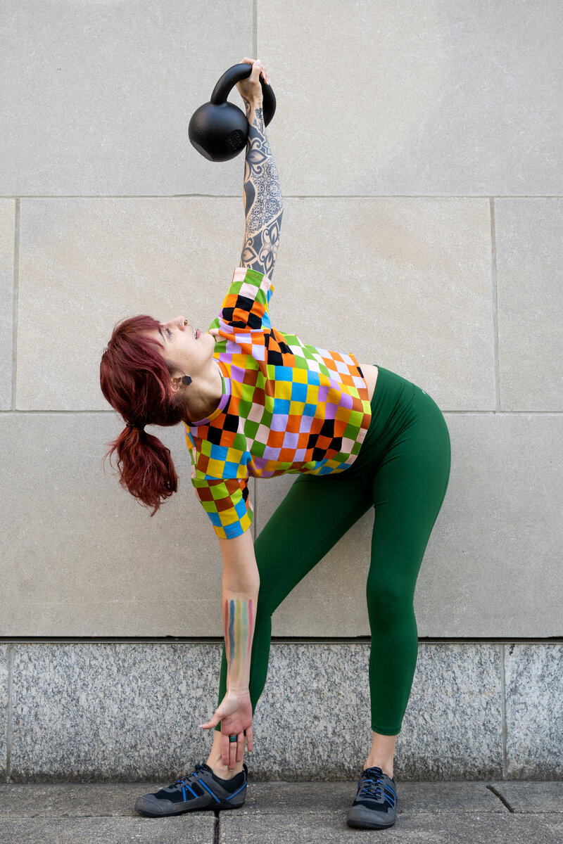 a woman in a rainbow checkered shirt does a kettlebell windmill