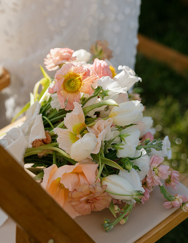 A bouquet of pastel flowers, including peach poppies and white tulips, rests on a wooden chair, with sunlight creating a soft, serene ambiance.