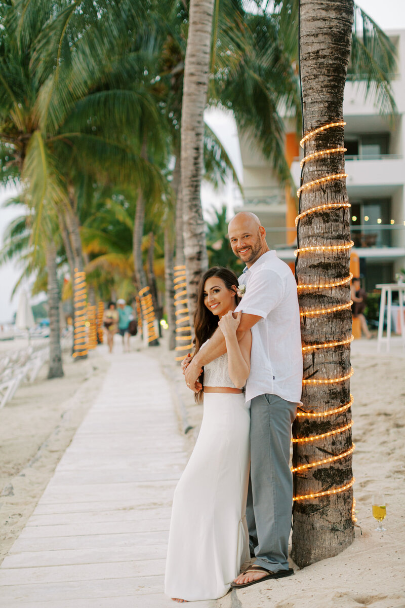 Couple poses against palm trees decorated with string lights along a tropical beachfront path.