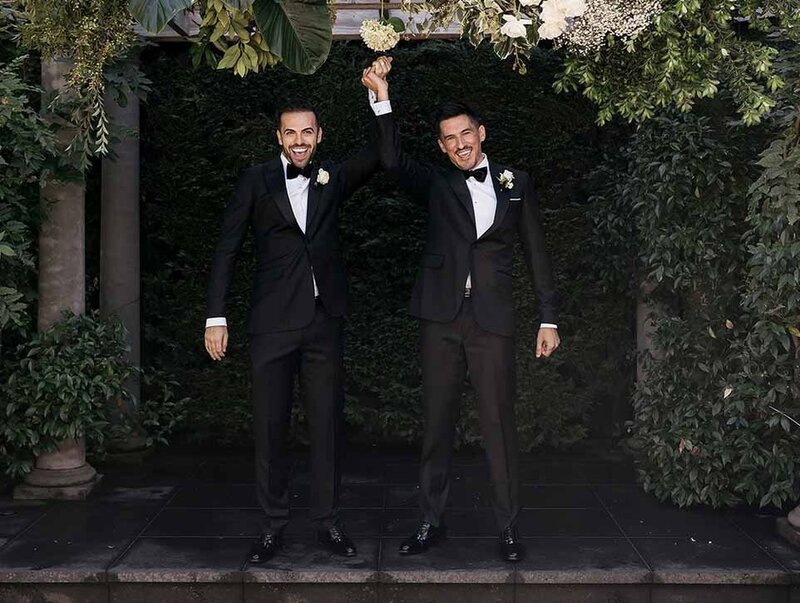 Two grooms in matching tuxedos celebrating under a floral arch at their outdoor wedding ceremony