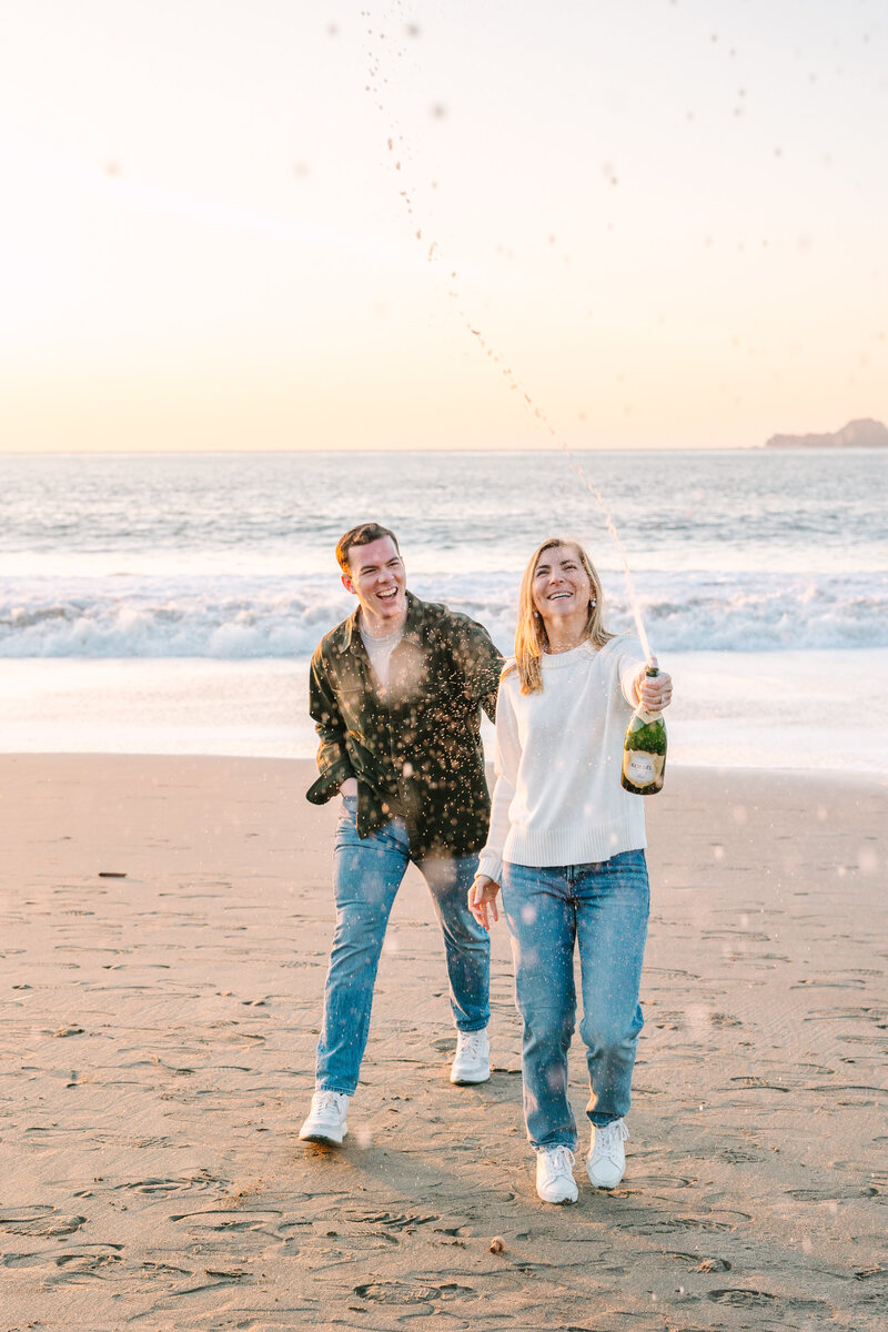 engaged couple on beach in san francisco  spraying a bottle of champagne while celebrating their engagement