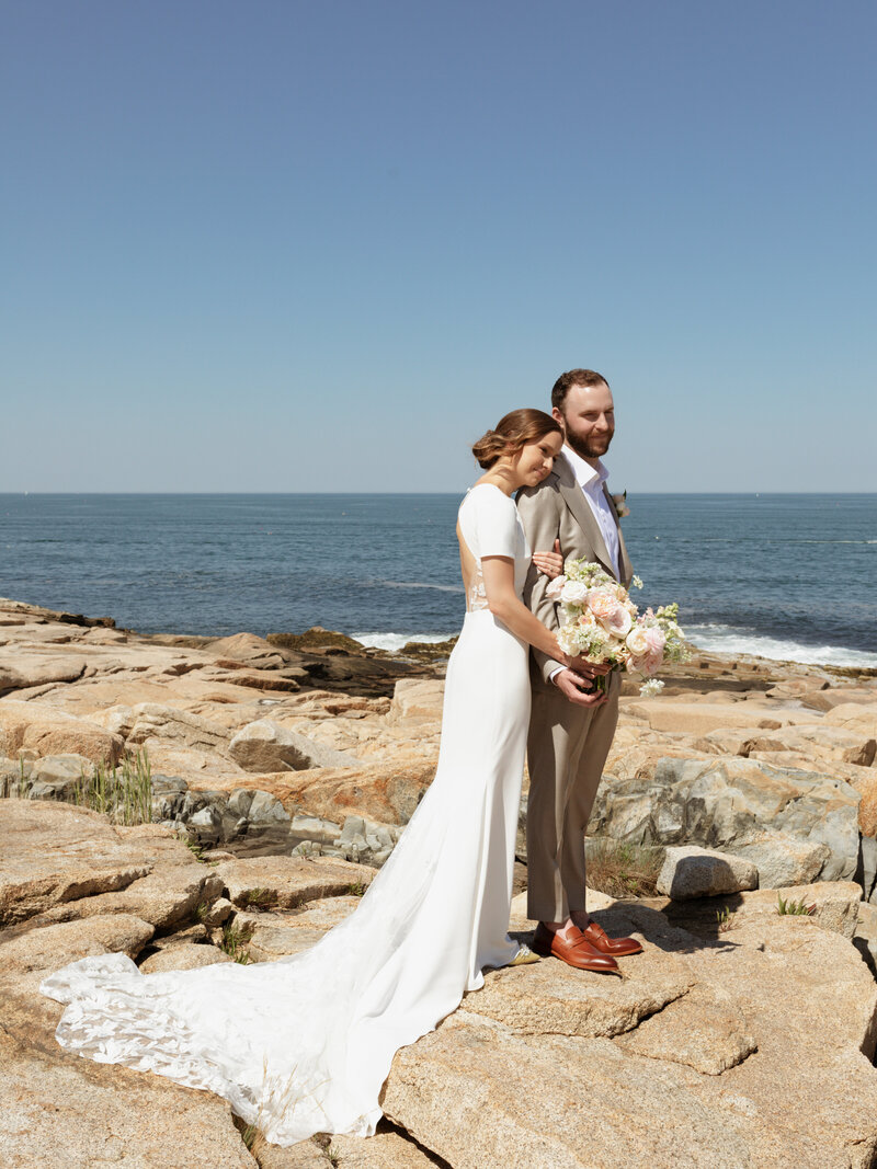 Bride and groom standing on rocks by the shore by Cape Cod wedding photographer Kelly Stevens.