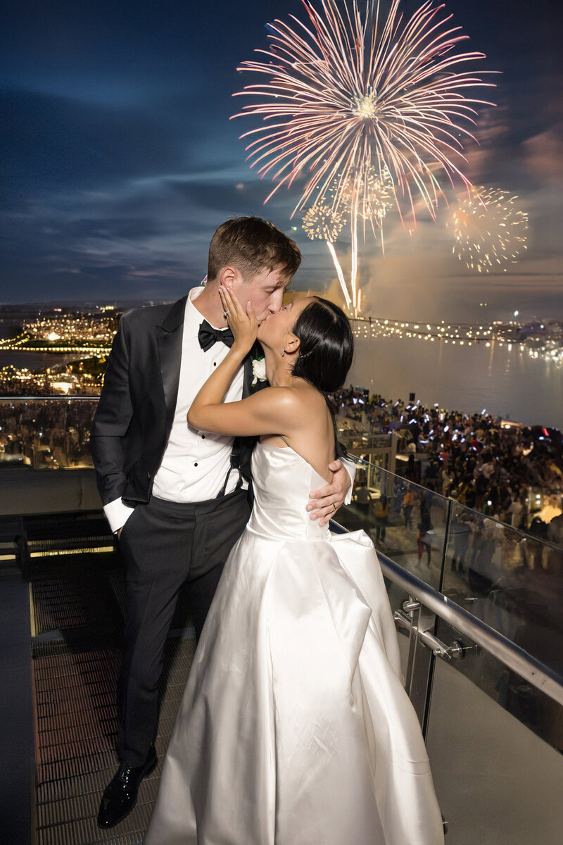 A couple in formal attire shares a kiss on a balcony overlooking a city waterfront. Fireworks light up the night sky above a crowd enjoying the celebration.