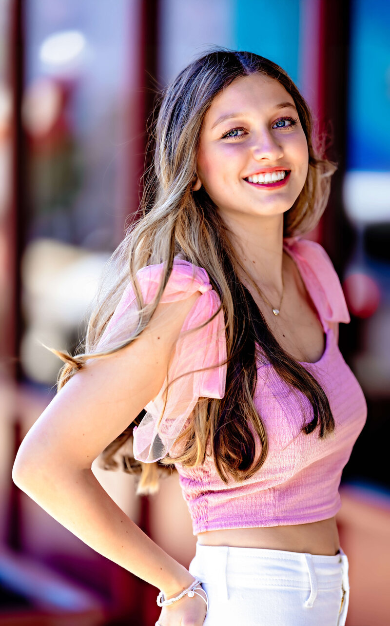 High school senior girl smiling in a pink top and white jeans during a colorful outdoor senior photo session in Winter Garden, Florida.