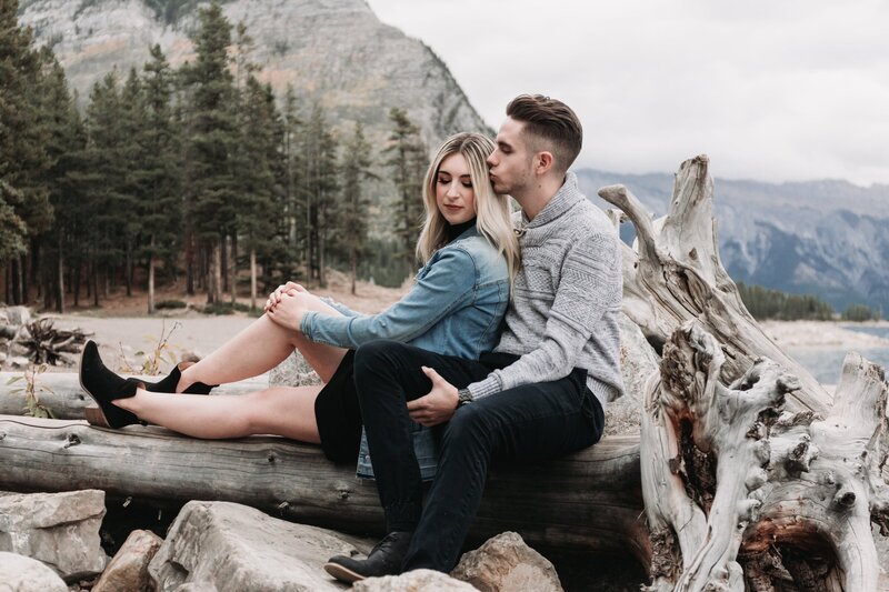 A young couple sit on driftwood on the shore of a mountain lake during their photography session. She has her hands clasped on one raised leg and is looking down and he's sitting beside her, kissing her on the side of her head.