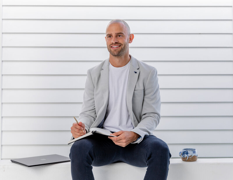 Jon Moses sitting with his laptop, notebook and coffee mug. in front of a white weatherboard wall