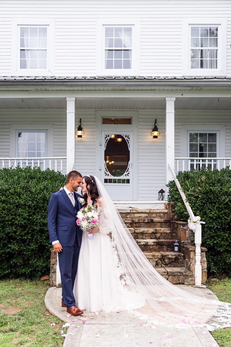 A bride and groom stand close together, smiling softly outside a white farmhouse with a stone staircase and classic porch. The bride’s long veil flows dramatically down the steps as she holds a bouquet of white and pink flowers, while the groom in a navy suit leans in toward her with affection.