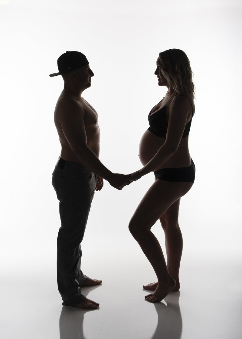 dark silhouette of male feamle couple facing each other holding hands in profile so mom to be's pregnant belly is showing and highlighted by the bright white background. Created in charlotte nc portrait studio by Insley Photography