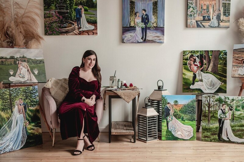 Debbie Bowman, wedding painter, sitting on a couch with several completed paintings around her