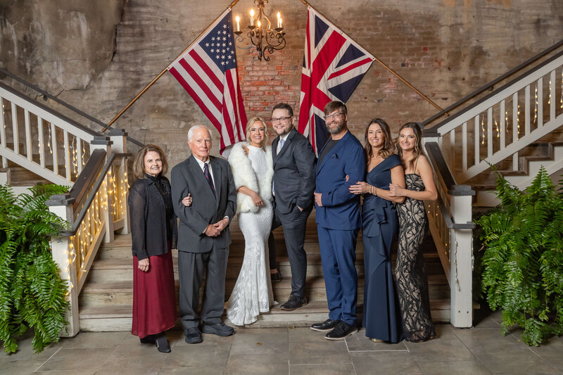 Seven people dressed formally stand on stairs decorated with string lights and ferns, with American and British flags behind them. The group smiles for the photo in front of a brick wall under a chandelier.