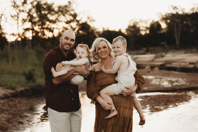 A couple stands outdoors near a body of water, both holding young children. The parents are smiling, and the sun is setting behind them. This serene moment perfectly captures the essence of family, making it an ideal scene for Christmas mini sessions NSW with an Illawarra newborn photographer.